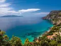 Turquoise water laps a rocky Tuscan coast with villas nestled among trees, under a blue sky with clouds.