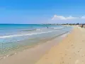 A wide, sandy beach with clear turquoise water and a few people swimming and sunbathing under a bright blue sky.