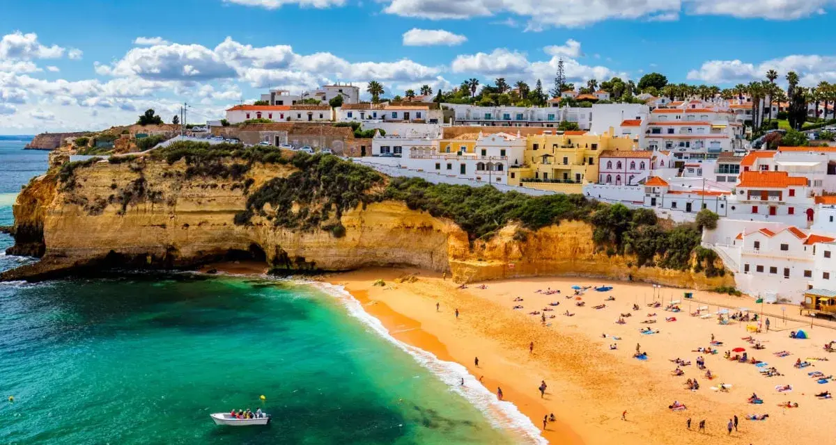 Coastal town with white buildings on cliffs overlooking a sandy beach and turquoise water.